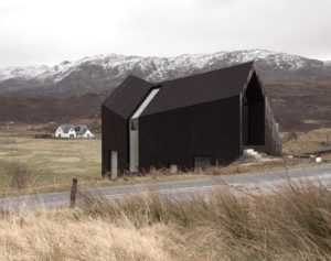 faceted black timber building next to a road with a snow peak mountain in the background