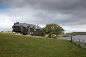 faceted dark timber house on a green grass area with a tree and loch at the end of the garden