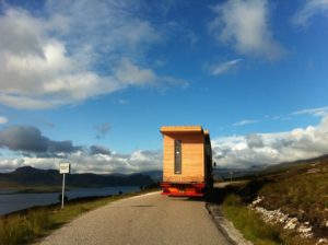 timber modular house on the back of a flatbed lorry on a single track road next to the sea and mountains