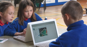 three children playing the my sust house game on laptops in a school gym