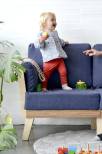 baby standing on a sofa with white walla nd grey rug and cheese plant