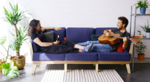 woman with a beer and a man with a guitar lying on a sofa facing each other surrounded by plats and a rug in front