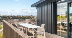 top floor of zinc clad building with timber balcony with modern timber railings, the decking has a table and chairs with distant views over Edinburgh